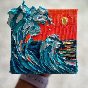 May include: A square canvas painting featuring thick, textured impasto oil paint. The artwork depicts a vibrant orange sky with a yellow sun above deep blue ocean waves with white highlights and splashes of bright orange paint.
