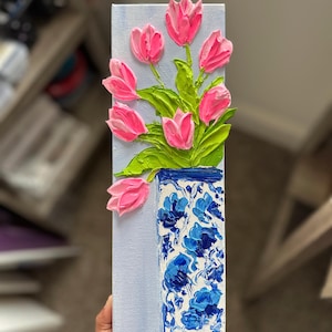 May include: A vertical painting of pink tulips in a blue and white vase. The tulips are textured and appear to be painted with thick brushstrokes. The vase has a floral design. The background is a light blue.