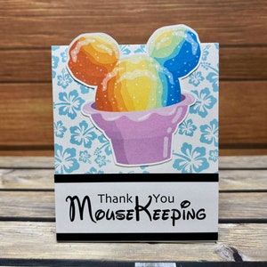 May include: A greeting card featuring a colorful, cartoon-style snow cone in the shape of Mickey Mouse ears. The card has a light blue background with white and blue floral patterns. The text reads "Thank You Mousekeeping."