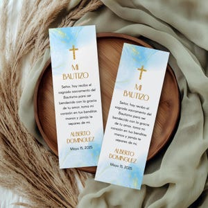 May include: Two blue and gold bookmark-style cards with a cross and the text "MI BAPTIZO" in gold. The text below reads "Señor, hoy recibo el sagrado sacramento del Bautismo para ser bendecido con la gracia de tu amor, toma mi corazón en tus benditas manos y jamás te separes de mi."  The name "ALBERTO DOMINGUEZ" and the date "Mayo 15, 2025" are printed at the bottom of each card.