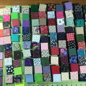 Könnte beinhalten: Eine Ansammlung kleiner, farbenfroher Stoffquadrate, die auf einer rosa und grünen Schneidematte angeordnet sind. Die Quadrate zeigen eine Vielzahl von Mustern und Farben, darunter florale, geometrische und abstrakte Designs. Die Stoffe sind wahrscheinlich für Quilting- oder andere Bastelprojekte bestimmt.
