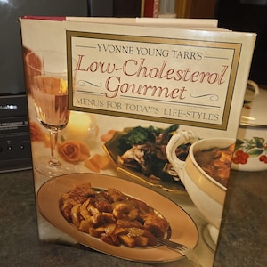 以下が含まれることがあります： 「Yvonne Young Tarr's Low-Cholesterol Gourmet: Menus for Today's Life-Styles」というタイトルのハードカバー料理本。表紙には、温かみのある背景を前に、盛り付けられた料理、ワイングラス、ボウルが写った写真が掲載されています。