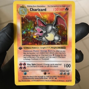 May include: A Charizard Pokémon card with a holographic background. The card features a gray and orange dragon with the text "Charizard" and "120 HP". The card also includes the text "Fire Spin" and "LV. 76 #6".