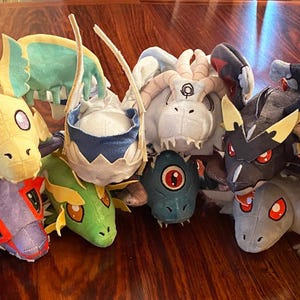 May include: A collection of 12 plush toys, each depicting a different dragon. The dragons are in various colours, including blue, green, yellow, and purple. They have different facial expressions and features, such as horns, wings, and scales.