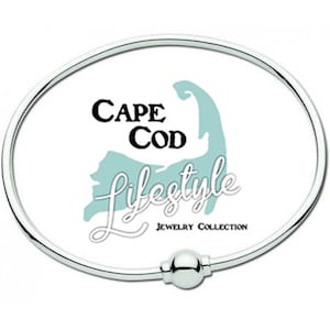 Pu&ograve; includere: Un braccialetto in argento con chiusura a sfera. Il braccialetto presenta uno sfondo bianco con un contorno blu di Cape Cod e il testo "Cape Cod Lifestyle Jewelry Collection".
