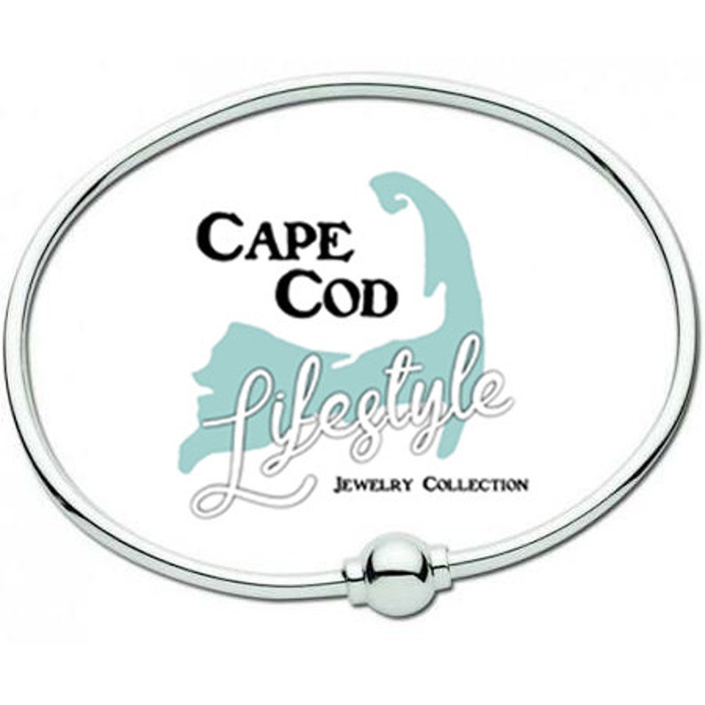 Pu&ograve; includere: Un braccialetto in argento con chiusura a sfera. Il braccialetto presenta uno sfondo bianco con un contorno blu di Cape Cod e il testo "Cape Cod Lifestyle Jewelry Collection".