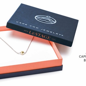Pu&ograve; includere: Una collana d'argento con un ciondolo a forma di perla dorata, presentata in una scatola blu navy con il testo "Cape Cod Jewelry" e "Lestage" stampato sul coperchio. La scatola ha un interno rosa corallo.