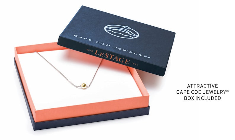 Pu&ograve; includere: Una collana d'argento con un ciondolo a forma di perla dorata, presentata in una scatola blu navy con il testo "Cape Cod Jewelry" e "Lestage" stampato sul coperchio. La scatola ha un interno rosa corallo.