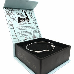 May include: A silver bangle bracelet with a nautical rope design, in a black box with a white interior. The box has a blue lid with a graphic of a lighthouse and the text "Cape Cod Lifestyle Jewelry Collection".