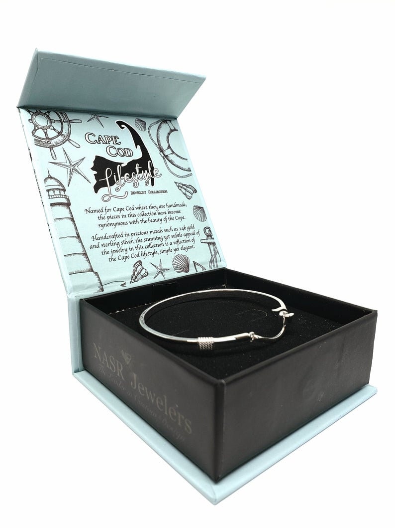 May include: A silver bangle bracelet with a nautical design, inside a blue and black box with the text "Cape Cod Lifestyle Jewelry Collection" and "Nasr Jewelers The Coastal Custom Design".