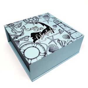 May include: A light blue box with a black and white illustration of a lighthouse, seashells, an anchor, and a life preserver. The box has the text "Cape Cod Lifestyle" printed on it.