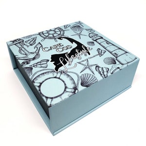 May include: A light blue box with a nautical theme. The box is decorated with a black and white illustration of a lighthouse, seashells, an anchor, a life preserver, and a compass. The text "Cape Cod Lifestyle" is printed on the box.