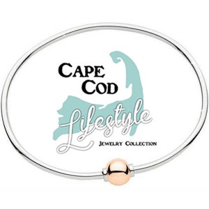 Pu&ograve; includere: Un braccialetto rigido in argento con un ciondolo a sfera in oro rosa. Il braccialetto presenta uno sfondo bianco con il testo "Cape Cod Lifestyle Jewelry Collection" e una grafica di Cape Cod.