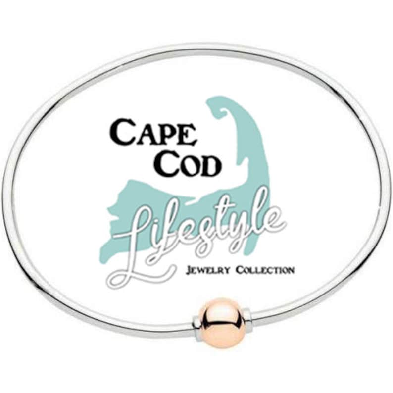 Pu&ograve; includere: Un braccialetto rigido in argento con un ciondolo a sfera in oro rosa. Il braccialetto presenta uno sfondo bianco con il testo "Cape Cod Lifestyle Jewelry Collection" e una grafica di Cape Cod.