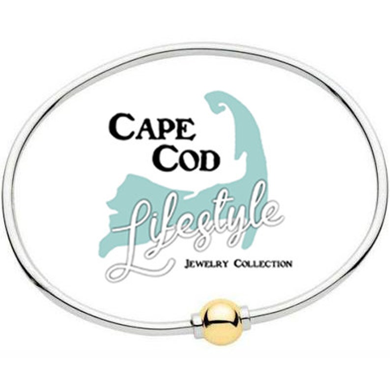 Pu&ograve; includere: Un braccialetto rigido in argento con un ciondolo a sfera dorata. Il braccialetto presenta uno sfondo bianco con un contorno blu di Cape Cod e il testo "Cape Cod Lifestyle Jewelry Collection".