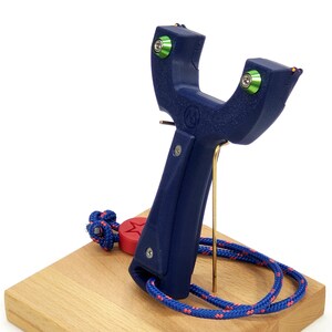 May include: A blue slingshot with green accents and a red star on the handle. The slingshot is on a wooden stand with a blue and red rope.
