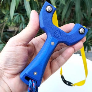 May include: A blue slingshot with a textured grip and a yellow band. The slingshot has a logo on the front and is held in a hand. The band is attached with metal fasteners. A red and blue rope is attached to the bottom.