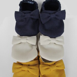 May include: Three pairs of baby shoes with bows. The top pair is navy blue, the middle pair is cream, and the bottom pair is mustard yellow.