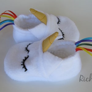 May include: White fleece baby booties with a unicorn design. The booties have a gold horn and rainbow tail.