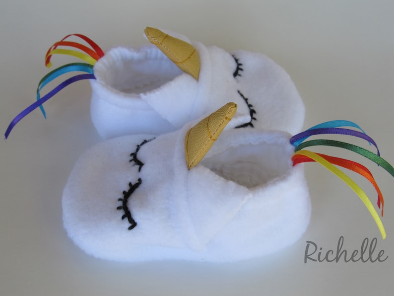 May include: White fleece baby booties with a unicorn design. The booties have a gold horn and rainbow tail.