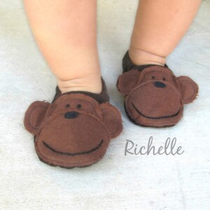May include: A pair of brown baby slippers with monkey faces. The slippers have black eyes and a stitched smile.