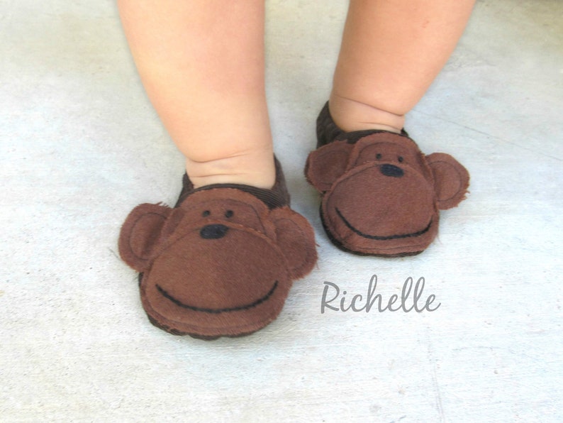 May include: A pair of brown baby slippers with monkey faces. The slippers have black eyes and a stitched smile.