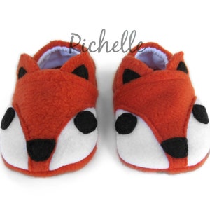 May include: A pair of orange fox-shaped baby booties with white and black felt accents. The booties are made of soft fleece and have a white lining.