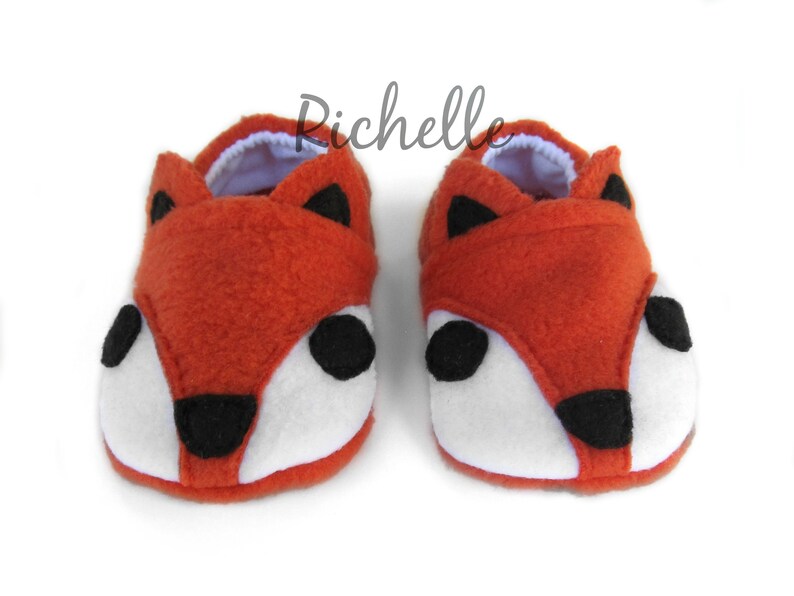 May include: A pair of orange fox-shaped baby booties with white and black felt accents. The booties are made of soft fleece and have a white lining.