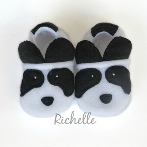 May include: A pair of gray and black panda-themed baby slippers. The slippers have black ears, eyes, and a nose.