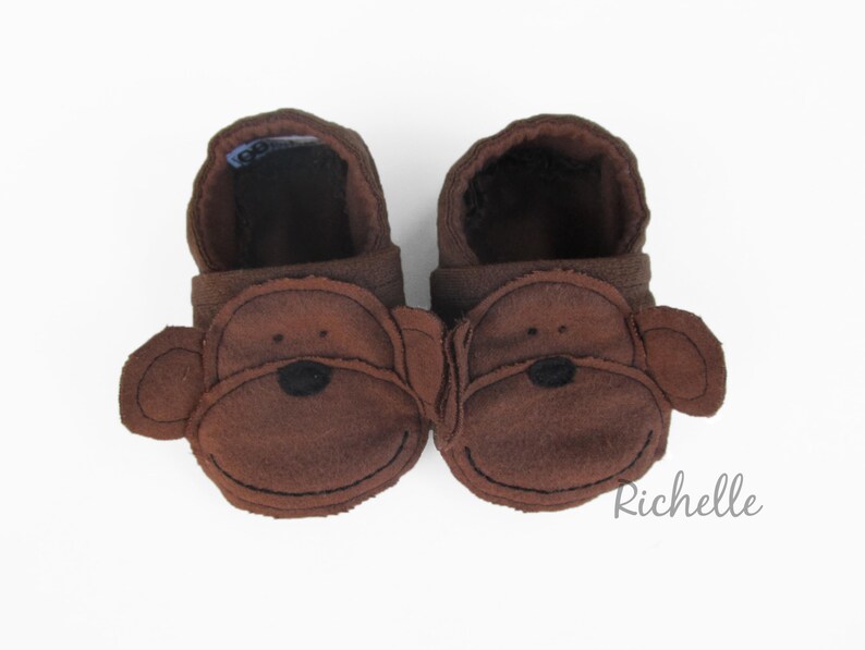 May include: A pair of brown baby booties with monkey faces. The booties are made of soft fabric and have a simple design.