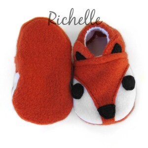 May include: A pair of orange fleece baby booties with a fox face design. The booties have black eyes, a black nose, and white fur around the eyes and mouth.