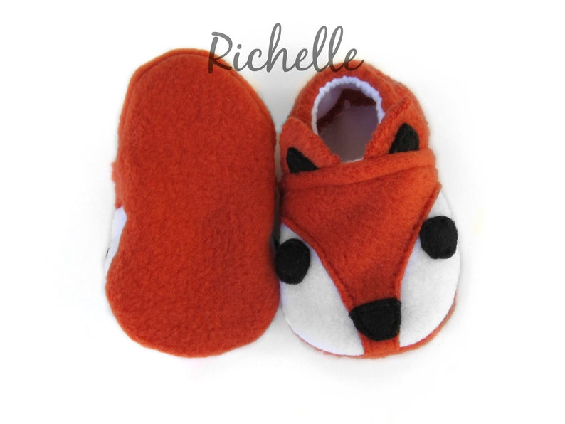 May include: A pair of orange fleece baby booties with a fox face design. The booties have black eyes, a black nose, and white fur around the eyes and mouth.