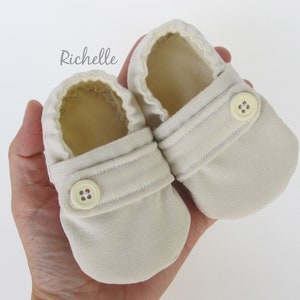 May include: A pair of beige baby booties with a button closure. The booties are made of a soft, textured fabric.