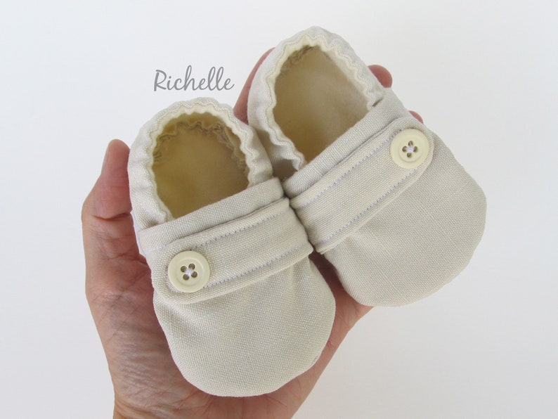May include: A pair of beige baby booties with a button closure. The booties are made of a soft, textured fabric.