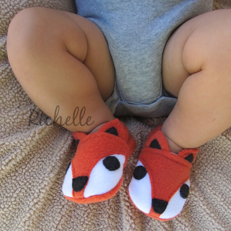 May include: A pair of orange baby booties with white and black fox faces. The booties are made of soft, fuzzy fabric.