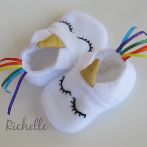 May include: White baby booties with a unicorn design. The booties have a gold horn, black embroidered eyes, and a rainbow ribbon tail.