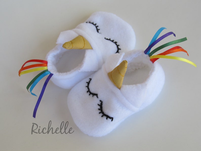 May include: White baby booties with a unicorn design. The booties have a gold horn, black embroidered eyes, and a rainbow ribbon tail.