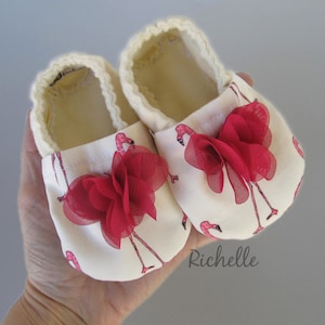 May include: A pair of white baby shoes with pink flamingo print and red fabric flower accents. The shoes are made of soft fabric and have a comfortable fit.