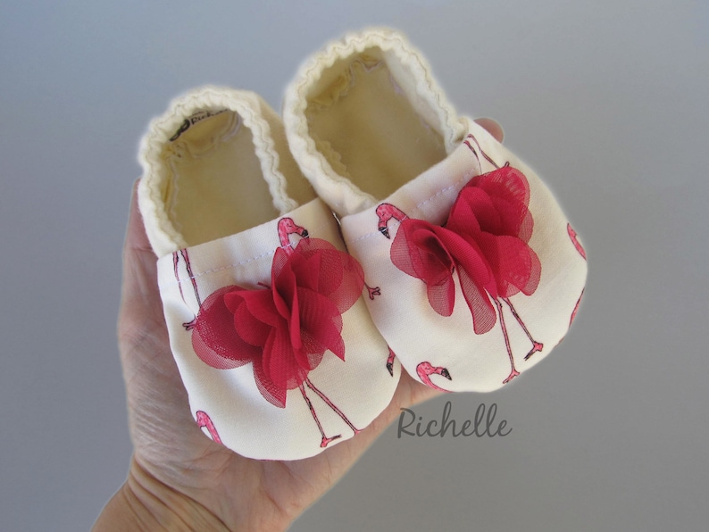 May include: A pair of white baby shoes with pink flamingo print and red fabric flower accents. The shoes are made of soft fabric and have a comfortable fit.