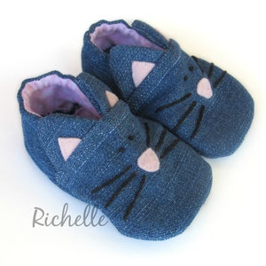 May include: A pair of blue denim baby shoes with a cat face design. The cat face features black whiskers and pink felt ears and nose.