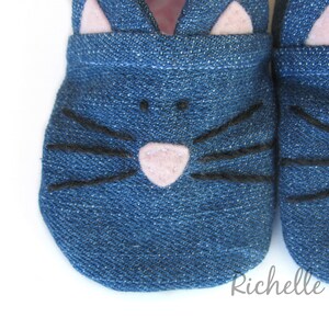 May include: A pair of blue denim baby shoes with a cat face design. The shoes have pink felt ears and a pink felt nose. The whiskers are black thread.