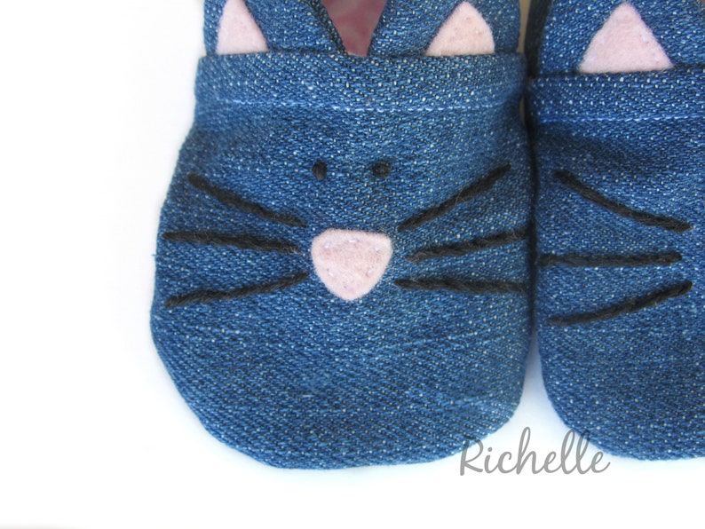 May include: A pair of blue denim baby shoes with a cat face design. The shoes have pink felt ears and a pink felt nose. The whiskers are black thread.