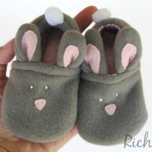 May include: A pair of gray baby booties shaped like bunnies, with pink inner ears, a pink nose, and white pom-poms. The booties are made of soft material and are designed to keep a baby's feet warm. The booties are held in a hand.