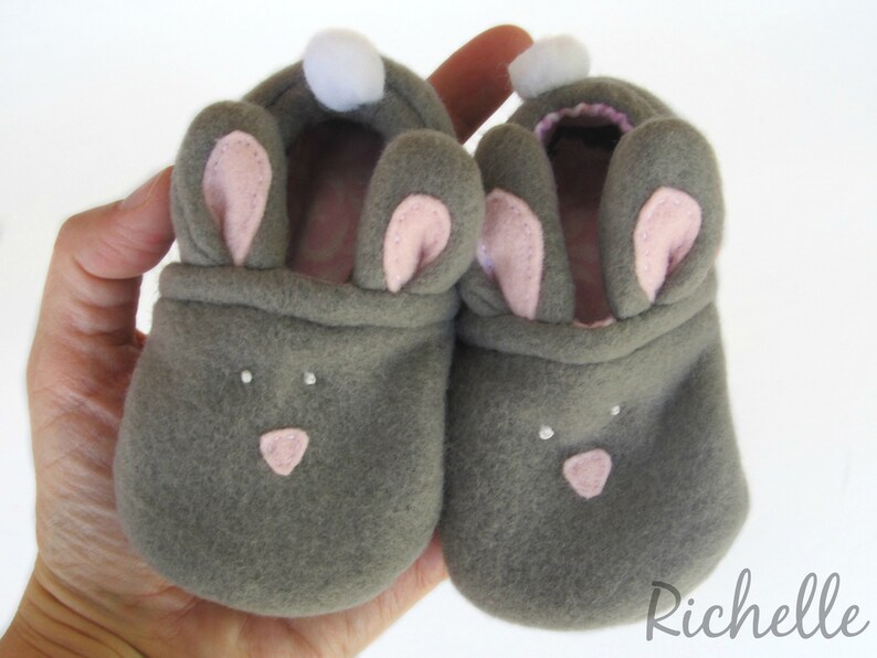 May include: A pair of gray baby booties shaped like bunnies, with pink inner ears, a pink nose, and white pom-poms. The booties are made of soft material and are designed to keep a baby's feet warm. The booties are held in a hand.