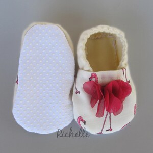 May include: A pair of white baby booties with a pink flamingo print and a red flower accent. The booties have a white dotted sole.