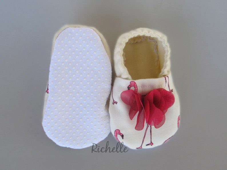 May include: A pair of white baby booties with a pink flamingo print and a red flower accent. The booties have a white dotted sole.