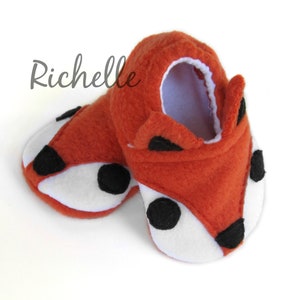 May include: A pair of orange fleece baby booties with black and white fox faces. The booties have a white lining and black felt accents for the eyes, nose, and ears.