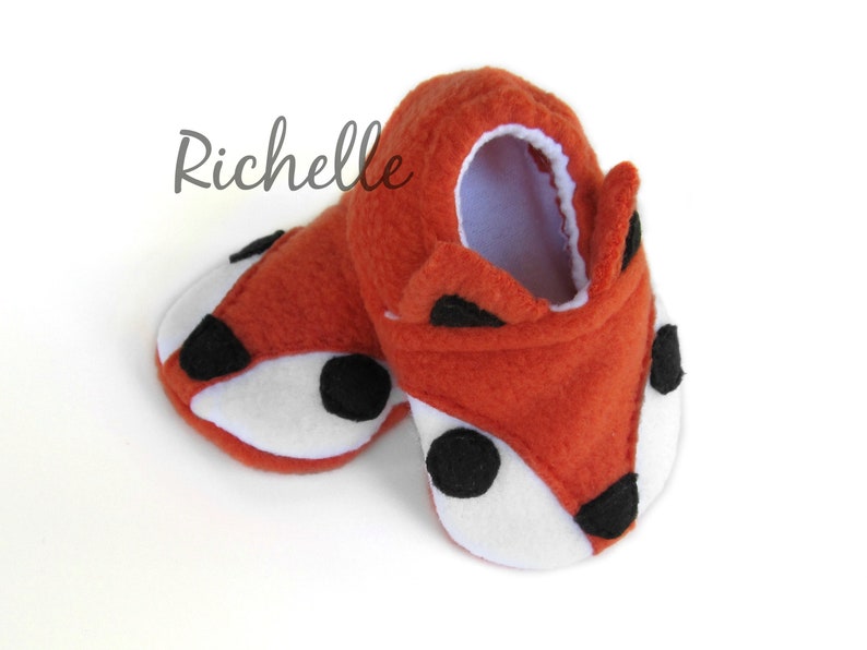 May include: A pair of orange fleece baby booties with black and white fox faces. The booties have a white lining and black felt accents for the eyes, nose, and ears.