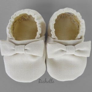 May include: A pair of off-white baby shoes with a bow on the front. The shoes are made of a soft, textured fabric.