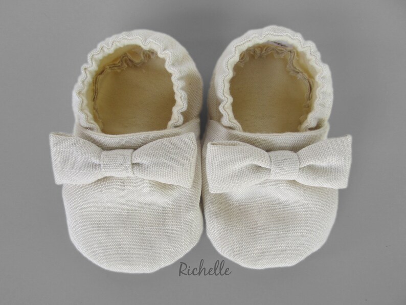 May include: A pair of off-white baby shoes with a bow on the front. The shoes are made of a soft, textured fabric.